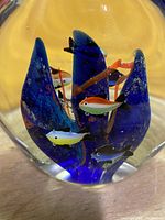 Close-up of fish figurine suspended inside blue glass coral formation within the clear glass body, showing detail of small colorful fish and bubbles.