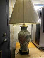 Full view of table lamp with white fabric shade and large hand-painted ceramic base showing floral design and brass hardware on wooden stand.