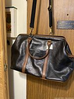 Front view of black and brown leather Fossil duffle bag hanging on door with handles and shoulder strap visible.