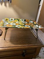 Side view of the foldable food tray with legs extended, placed on top of a wooden surface showing the colorful floral daisy design.