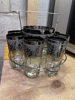 Set of six tall glasses with silver grape and vine design in a silver metal circular basket holder with handle on wooden surface against concrete wall background.