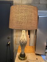 Side view of the full table lamp showing the terracotta clay base with geometric patterns and beige textured fabric cylindrical lampshade, set on a brass circular base.