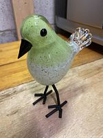Front and side view of light green and white Murano glass bird with black beak and metal legs on wooden surface.