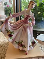 Full view of porcelain lady figurine in pink dress with floral patterns standing on flat surface near window.