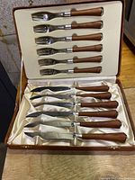 Open case showing six wooden-handled forks fitted in satin-lined top case section and six wooden-handled steak knives in bottom section.