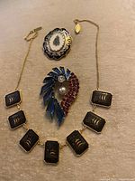 Photo showing vintage necklace with linked rectangular gold-tone plates with bird engravings and two brooches: oval floral motif and rhinestone fan shape
