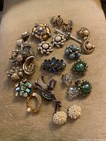 Photo showing all vintage brooches and earrings laid out on a beige surface. Various designs include pearl clusters, colored rhinestones, enamel flower brooch, and gold-tone pieces.