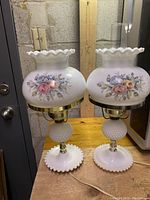 Front view of both vintage milk glass lamps showing floral hand-painted shades and hobnail bases, placed on wooden surface.