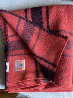 Folded red wool blanket showing label of Plessis Woollen Mills Ltd. and part of the plaid design along with black whipstitch edging.