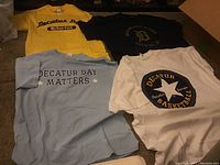 Four Decatur Day T-shirts laid out in box showing different design and colors: yellow with McKoy Park, black with Old English B and Decatur text, light blue with 'Decatur Day Matters', white with Decatur Basketball and star logo.