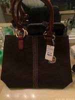 Front view of a brown textured leather-like purse with double handles and a gold-tone medallion charm, showing a new tag attached to the handle.