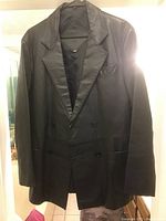 Front view of the men's black leather jacket hanging, showing double-breasted front with four buttons, peak lapels, and two lower front pockets plus one chest pocket.