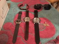 Photo showing three men's wristwatches with different band styles and two pairs of sunglasses plus a pin that says "REAL".
