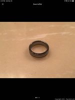 Photo of the tungsten carbide men's wedding band on a beige background showing front and side. The ring is metallic gray with three horizontal grooves.