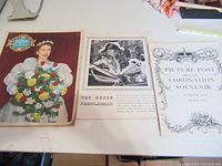 Shows three items laid flat: full-color coronation magazine with Queen Elizabeth II on cover holding bouquet, a black and white printed coronation page, and an ornate special coronation souvenir cover page