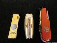 Three items laid out on black cloth: two vintage lighters, one gold-tone Taurus emblem lighter and one silver-tone lighter with wear, plus red multi tool pocket knife with star emblem.