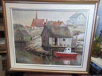 Full view of the framed oil painting depicting a fishing scene with a red and white fishing boat near wooden shacks by the water, houses in the background.
