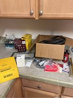 Photo showing cardboard box with black jumper cables, metal license plate frames, yellow box, automotive fluid container, and small items on kitchen counter.