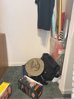 Photo showing gift wrap rolls, wide-brim hat, black wheeled bag, and other small boxed items on carpeted floor near closet wall.