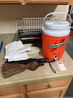 Photo showing orange Gatorade drink dispenser, metal breadbox, white oven mitts, and two umbrellas on countertop.