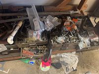Wide view of boxes filled with assorted hardware pieces including nuts, bolts, washers, metal brackets, some in clear plastic bags, hand tools, and pipes.