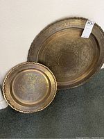 Photo showing two metal wall platters leaning against wall, one larger with simple border, one smaller with scalloped edge