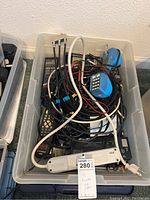 Photo of plastic bin containing various remote controls, tangled cables, and blue phone line tester devices.