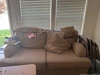 Full front view of beige sofa with five pillows, placed near window blinds and accompanied by stacked folding chairs.