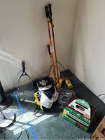Photo showing a variety of sport equipment including ski poles, tennis rackets, dumbbells, tennis balls, and hats gathered on a carpeted floor corner.