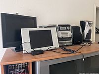 Photo showing Sharper Image CD player, AIWA stereo system with two black speakers, two Gem monitors in black and silver, and Sylvania 13-inch LCD TV below the shelf.