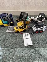 Lot overview showing four miniature toy construction vehicles and a small accessory bag on carpeted floor