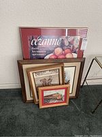 Four framed pieces of wall art stacked leaning against a wall, showing visible details of the largest framed print with 'Cézanne' text and fruit artwork.