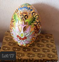 Front view of the cloisonné egg showing detailed lotus flower and swirling gold patterns. Egg rests on decorative box featuring brown and gold ornate design.