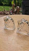 Pair of clear glass horse head bookends shown side by side on wooden surface with outdoor background.