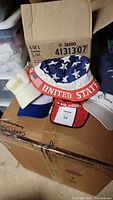 Photo of the six hats in a cardboard box, showing a USA bucket hat and Tommy Hilfiger cap