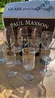 Photo showing eight of the wine glasses arranged in front of a Paul Masson brandy box, varying in size and base color including clear, green, blue, and frosted styles.