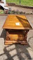 Front and top view of wooden side table outdoors, showing tabletop, drawer with handle, turned legs, and base molding