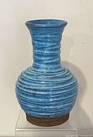 Front view of blue textured ribbed vase with wide rounded body, tall neck, and brown manganese base on transparent stand.