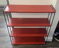 Front view of vintage metal 4-tier shelf with red shelves and black frame against white wall.