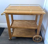 Angle view showing the three shelves, brass railing on the top shelf, wooden structure, large wooden wheel on one side and small caster wheels on the other side.