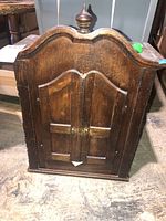 Front view of the small wooden cabinet showing curved top and two paneled doors with finial