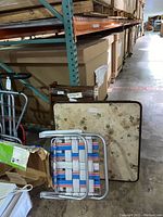 Card table and aluminum outdoor chair folded and stacked against shelves in a warehouse.