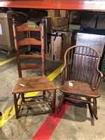 Photo showing the two chairs side by side, on a warehouse floor. Left chair is ladder back with cane woven seat, right chair is wooden rocking chair with multiple thin vertical slats and curved armrests.