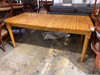 Full view of the butcher block dining table showing rectangular tabletop with butcher block pattern, four rounded and tapered legs, sitting on a concrete floor, surrounded by various chairs and furniture.