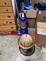 Photo showing blue and red Hoover Fusion vacuum cleaner standing on carpet near wooden dresser, with vintage-style light-up globe positioned on the floor.