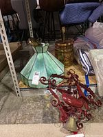View of three items: green stained glass Tiffany style lamp shade, barrel shaped wood and brass table lamp, red painted metal bell door chime.