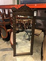 Front view of the wood framed decorative wall mirror showing carved top and sides, and clear mirror glass reflecting warehouse surroundings.