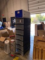 Stack of eight plastic stackable storage bins with mostly black bodies and transparent drawers, one bin with a white body and blue front drawer, shown in a cluttered warehouse environment.
