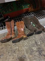 Photo showing brown leather boots with side zipper and chunky heel next to tall grey suede boots on a concrete floor.
