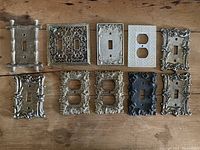 Top view of all 10 vintage decorative switch plate covers arranged on wooden surface, showing variety of styles and finishes.